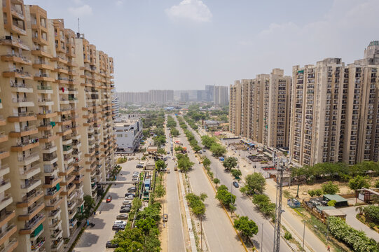 Aerial view of modern high-rise residential apartment complexes flanking a wide road with parked cars and green trees in Greater Noida, Uttar Pradesh, India.