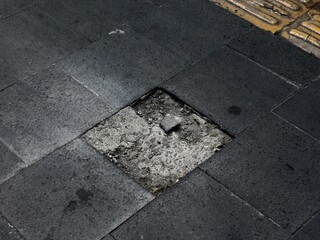 Fototapeta premium Missing black sidewalk tile exposing broken concrete and debris, a tripping hazard on urban pavement