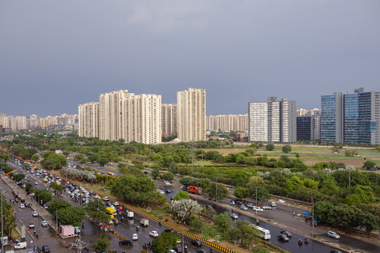 Aerial view of modern high-rise residential and office buildings overlooking a busy multi-lane road with heavy traffic, surrounded by lush green trees in Noida, Uttar Pradesh, India.
