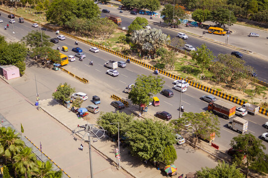 Aerial view of a busy multi-lane road with cars, trucks, and buses navigating through the urban landscape, surrounded by green trees in Noida, Uttar Pradesh, India.