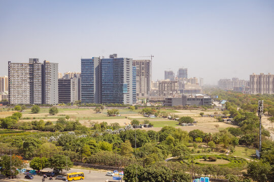 Aerial view of modern high-rise buildings and office complexes rising above green fields and urban parkland in Noida, Uttar Pradesh, India.