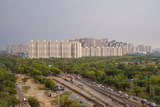 Aerial view of modern residential high-rise apartment complexes surrounded by lush green trees and a busy road in Noida, Uttar Pradesh, India.
