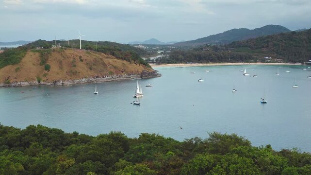 Nai Harn Beach in Phuket, Thailand, numerous sailboats and yachts anchored in peaceful bay, surrounded by lush green hills and a prominent wind turbine. Travel destination. Aerial view drone flight