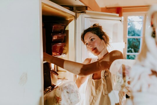 A young woman placing food containers into an open refrigerator with a soft glowing interior for kitchen organization and meal prep concepts