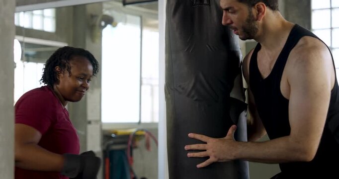 Plus size African American woman practices boxing beside heavy bag as male trainer coaches stance and punch timing in modern gym, building cardio fitness, strength, teamwork, motivation, endurance.