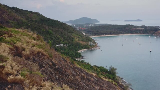 Aerial view capturing the rugged coastline of Phuket island, featuring mountains, lush greenery, and the tranquil bay of Nai Harn beach dotted with boats, under a soft sky