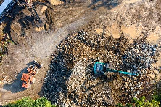 Aerial view of heavy machinery including an excavator and a loader working at a stone quarry with large piles of rocks and muddy ground in Karanglewas, Central Java, Indonesia.