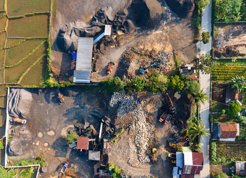 Aerial view of the Babakan stone crushing site with piles of gravel, heavy machinery, and trucks, near agricultural fields in Karanglewas, Central Java, Indonesia.