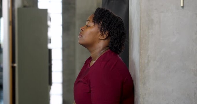 African American plus size woman pauses after boxing session, glove near chin while lifting face upward beside gym pillar. Curvy female athlete regains breath, wellness, confidence, recovery routine.