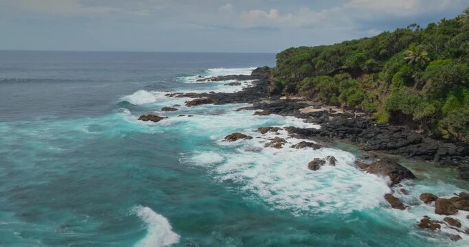 Aerial view drone flight over turquoise ocean waves crashing against the rugged volcanic rock coastline and a lush green tropical forest on Tanna Island, Vanuatu. Natural travel background