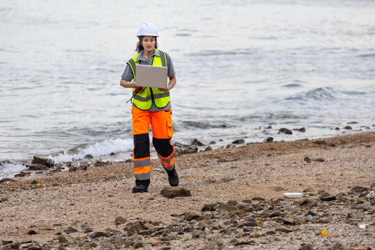 Female Environmental Scientist Conducting Beach Cleanup Survey, Engineer Using Laptop for Coastal Area Assessment, Worker in Safety Vest and Hard Hat Documenting Outdoor Site