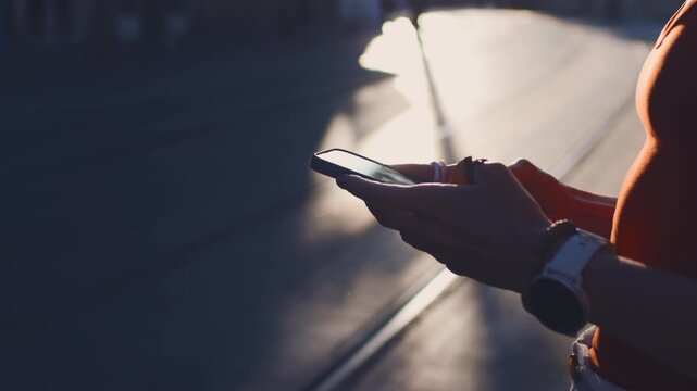 Close-up: a young woman holding a smartphone and typing a message. She is using the phone outdoors against a cityscape background. Urban lifestyle.