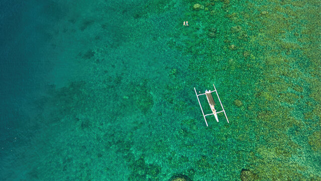 Aerial view of a traditional outrigger boat floating on clear turquoise water over a vibrant coral reef with two people swimming nearby in Kecamatan Abang, Bali, Indonesia.