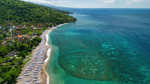 Aerial view of Bintang Beach Villa with traditional outrigger boats on the sandy beach, lush green hills, and turquoise coral reef waters in Kecamatan Abang, Bali, Indonesia.