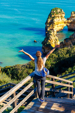 Happy female hiker with backpack standing on wooden stairs looking at ocean cliffs in Algarve Portugal