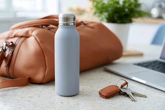 Gray water bottle stands on a clean countertop. Brown leather bag rests beside it. Keys lie near a laptop, suggesting travel or daily commute. Soft light illuminates a calm, organized workspace