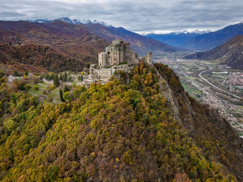 Aerial view of Sacra di San Michele perched on a rocky peak surrounded by vibrant autumn forests and the snow-capped Alps, Sant'Ambrogio di Torino, Piemonte, Italy.