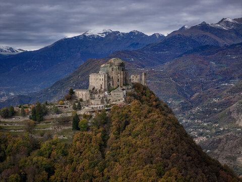 Aerial view of the historic Sacra di San Michele abbey perched on Mount Pirchiriano, surrounded by dense autumn forests and Alpine peaks in Sant'Ambrogio di Torino, Piemonte, Italy.