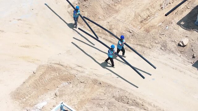 Three workers carrying long black pipes at a construction site on sandy terrain