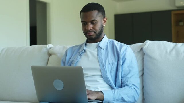A focused young African American man works from home, typing on his laptop while sitting comfortably on a modern white sofa. He occasionally pauses, gazing thoughtfully into the distance, perhaps cons