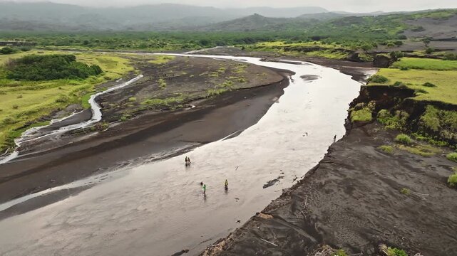 Winding river with dark volcanic ash and green vegetation, flowing through the Port Resolution area of Tanna Island, with people wading in the shallow water. Aerial view drone flight panorama