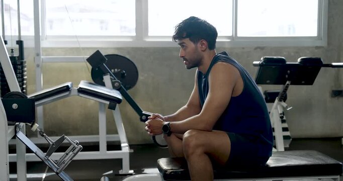 Young Asian man rests on gym bench after cable row exercise, leaning forward while holding handle and recovering between sets, solo arm strength routine, recovery, wellness, healthy active lifestyle.