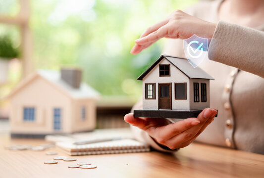 Home insurance and property protection concept. Woman hands holding miniature house model with secure shield checkmark icon.