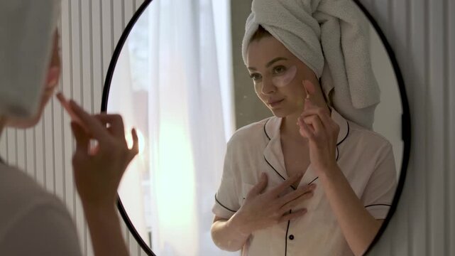 Young woman with a towel wrapped around her hair using a rose quartz facial roller while wearing under eye patches in front of a mirror. Home skincare, beauty treatment and self care wellness concept