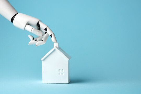 A white robotic hand touches a small white house model on a light blue background, symbolizing smart home technology, real estate, AI automation and future innovation.