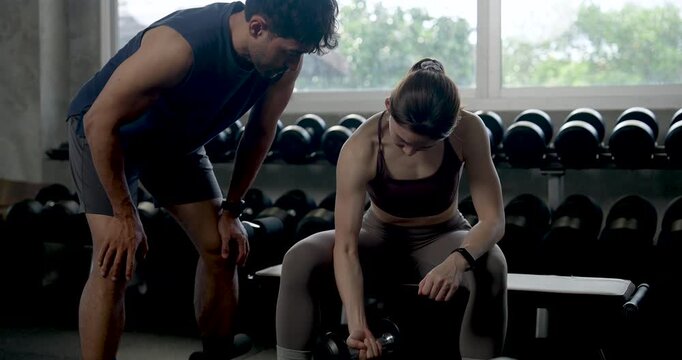 Young Asian man bends beside woman in modern gym, pointing at dumbbell while guiding seated curl form during partner workout, coaching strength training, support, motivation, healthy teamwork