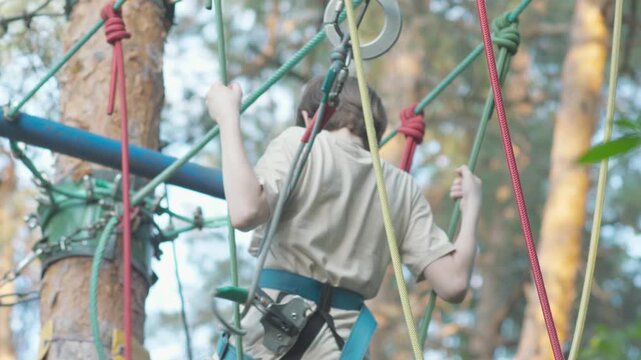 A boy in safety harness carefully navigates a challenging section in a rope adventure park. The child holds onto ropes and overcomes obstacles at a height among green trees.