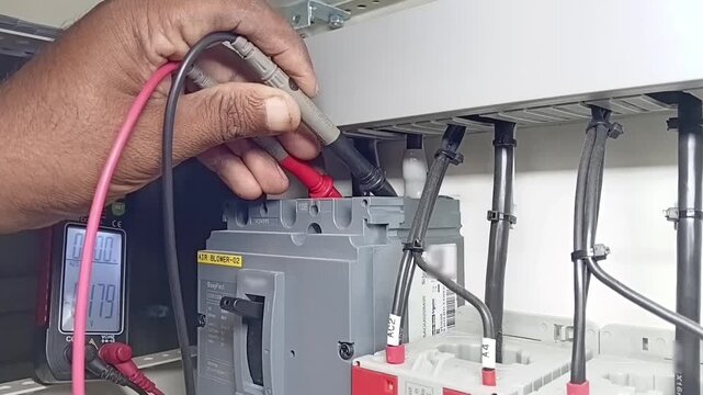 Electrician using digital multimeter probes to check voltage between L2 and L3 phases on industrial MCCB breaker inside electrical panel
