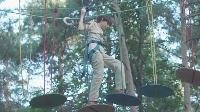 A boy wearing a safety harness overcomes a challenging stage in a rope park. The child carefully steps along suspended colorful discs at a height among a pine forest