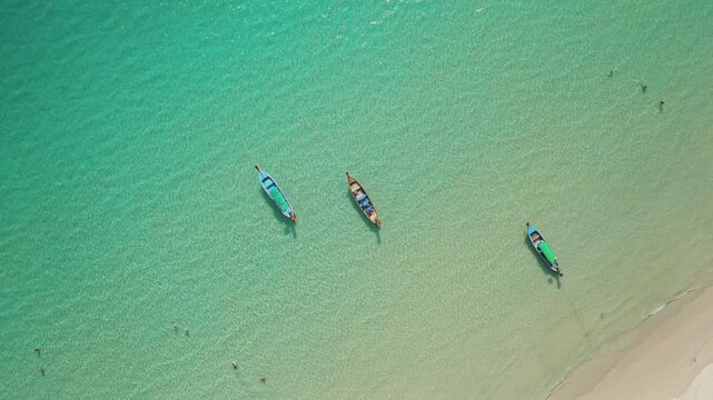 Longtail boats floating on the clear turquoise water of the Andaman Sea, with a few people swimming near the white sand beach, creating a peaceful tropical travel scene. Aerial top down drone shot
