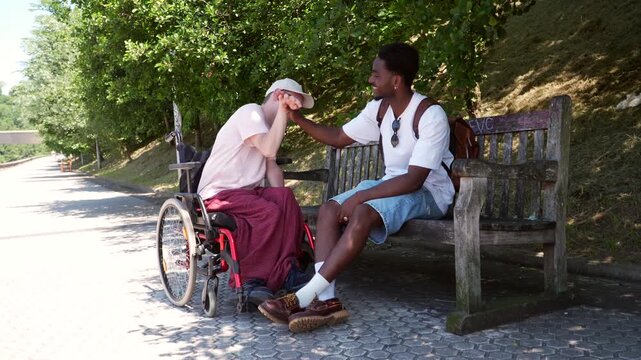 Two young diverse friends enjoying a sunny day outdoors, with one man in a wheelchair and the other sitting on a park bench, sharing a laugh and a special moment of connection and support