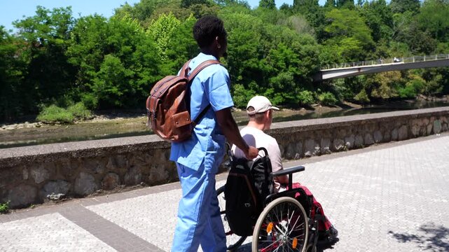 Young african american caregiver assisting an elderly disabled caucasian man in a wheelchair during a walk along the river in a sunny city park, providing essential support and companionship