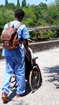 Young black caregiver in blue scrubs pushing an elderly disabled man in a wheelchair. Compassionate healthcare assistant providing support and walking with a senior patient in a park on a sunny day
