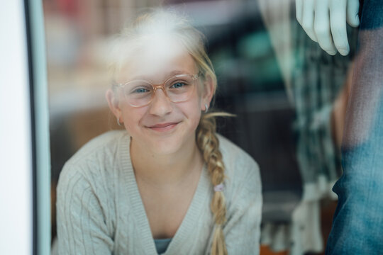 Cheerful child wearing eyeglasses gazing through the display window of a fashion boutique while observing the lively street outside.