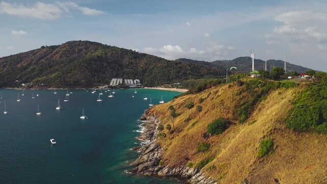 Tropical bay at Nai Harn Beach with numerous yachts anchored in turquoise water, a paraglider soaring above cliffs, and a wind turbine on Promthep Cape, Phuket, Thailand. Aerial view drone flight