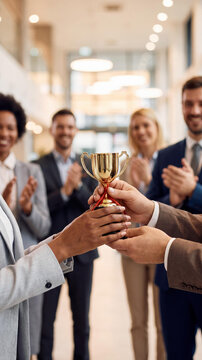 Close-up of a Black woman and Caucasian man holding a golden trophy cup. A diverse group of blurred business colleagues in suits applaud and celebrate their success in a bright office hall.