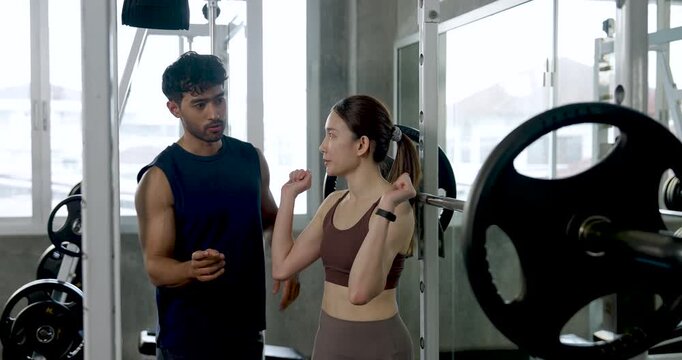 Young Asian couple learning barbell squat setup in modern gym, man adjusts rack area and guides woman near weight bar, partner workout with support, motivation, strength training, wellness goals.