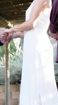 Vertical video: Tilting camera up, woman in white dress holding man's hands during garden ceremony