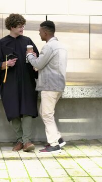 Vertical video: Walking friend handing cup to grad in gown yellow tassel at sunny plaza celebrating