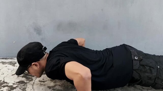 A young man in black sportswear performs push-ups outdoors on a concrete surface, demonstrating strength, endurance, and fitness training against a simple gray wall background