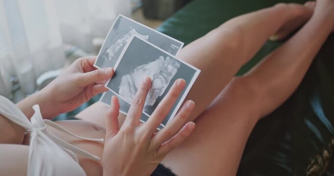 Pregnant woman holding and looking at baby ultrasound sonogram photos at home.