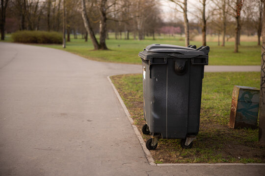 Black waste bin positioned on a paved pathway in a park, surrounded by trees and grassy areas, with a clear view of the landscape in the background