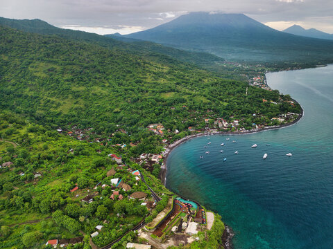 Aerial view of Jemeluk Beach Bungalows nestled among lush green hills along the black sand coast with Mount Agung in the background, Kecamatan Abang, Bali, Indonesia.