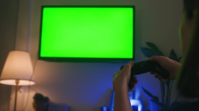Rear view of person holding a game controller while looking at television with green chroma key screen. Gaming, entertainment, streaming, smart TV and home leisure concept. High quality 4k footage