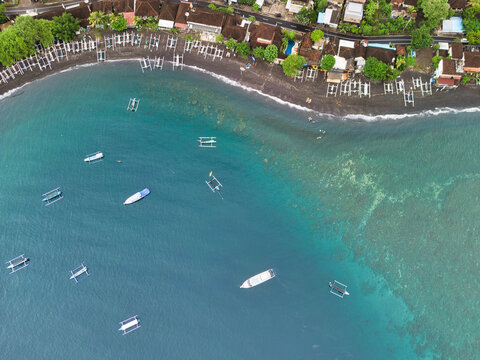 Aerial view of Jemeluk Beach Bungalows with traditional outrigger boats floating on turquoise water near the black sand coastline in Kecamatan Abang, Bali, Indonesia.