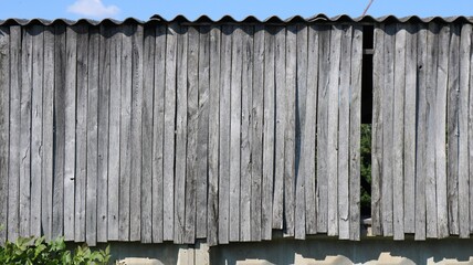 An old wooden fence wall of gray vertical boards. The faded wood is cracked and grained with age. Above, the corrugated slate roof of a barn is visible, and below, a concrete foundation and greenery. © Skyliz